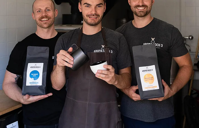 Drei junge Männer stheen nebneinadner in einer Kafferösterei. Links ein Herr mit kurzen Haaren und Bart, in der Hand eine Packung Kaffee. In der Mitte ein Mann mit braunen kurzen Haaren und Bart in der Hand eine Tasse und eine Kaffeekanne. Rechts ein Mann auch mit braunen kurzen Haaren und einem Bart, in der Hand auch eine Packung Kaffee.